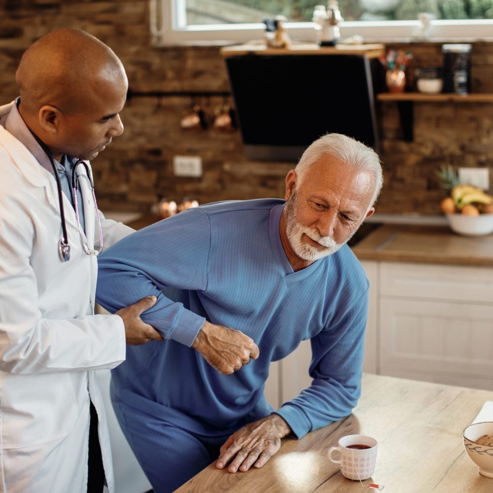 African American doctor helping senior man to get up from the chair during home visit.