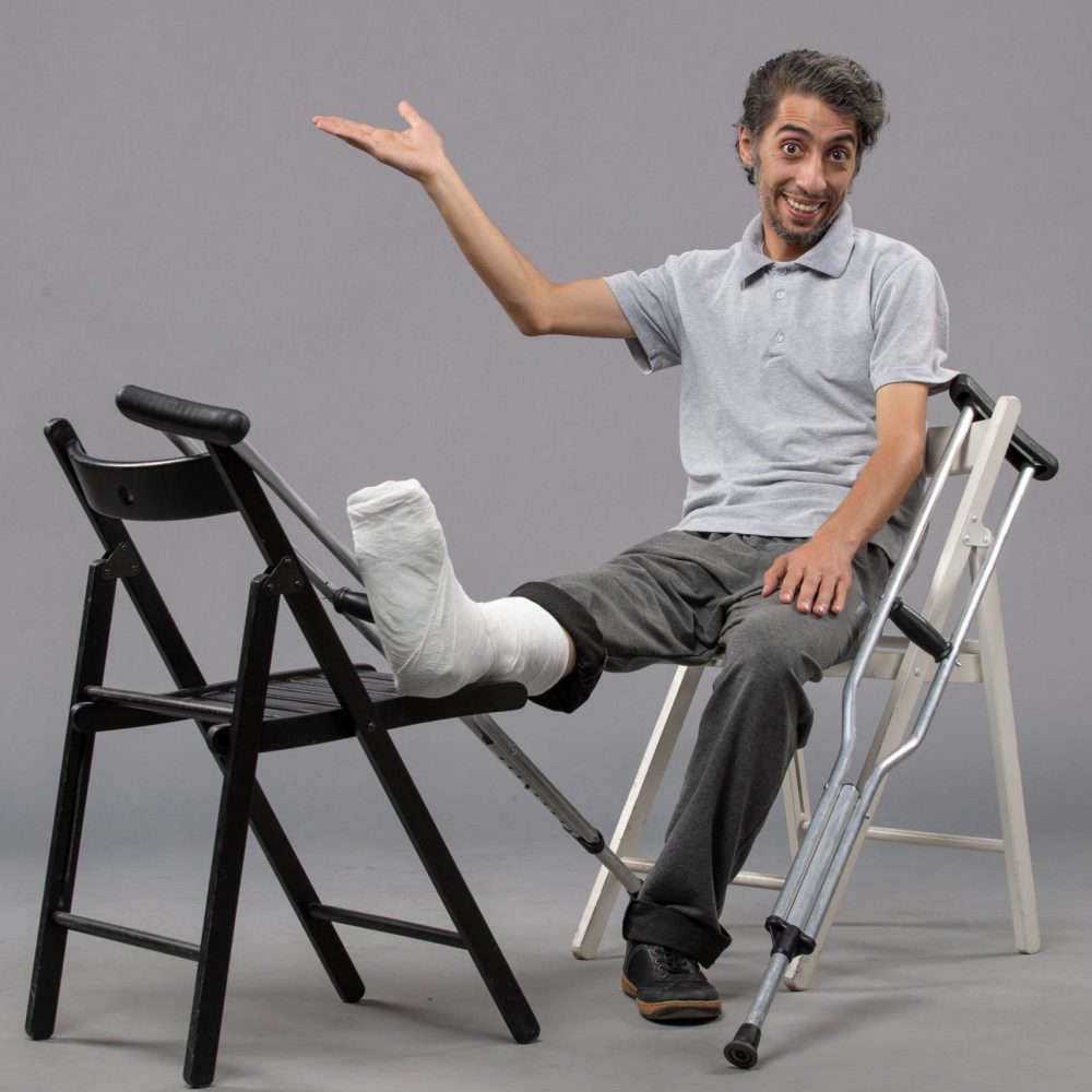 front-view-young-male-sitting-with-broken-foot-crutches-grey-wall-accident-broken-twist-foot-pain-leg