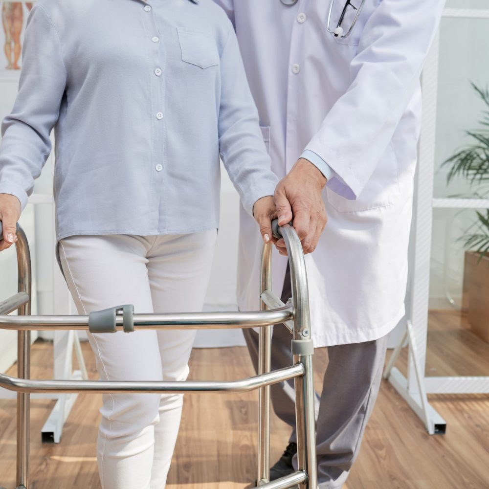 Close-up shot of unrecognizable physiotherapist explaining patient how to use front-wheeled walker, interior of modern office on background